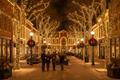 Festive main street at night with warm white seasonal lighting on trees, building facades, and lamp posts